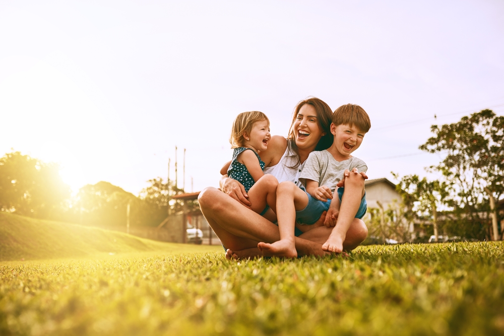 A happy mother sitting on the grass with her young daughter and son, all laughing and smiling together under a warm sunset—capturing the joyful, family-centered spirit of Little Koi Pediatric Dentistry