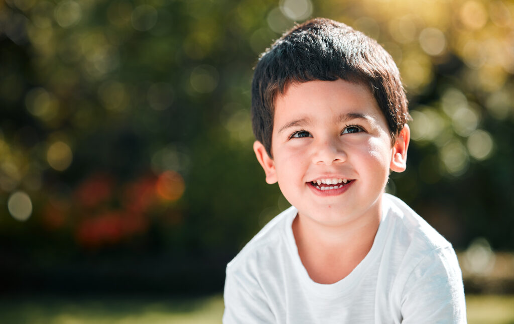 A cheerful young boy wearing a white t-shirt smiles brightly while sitting outside in natural light, conveying warmth, trust, and the joyful spirit of pediatric care – Little Koi Pediatric Dentistry